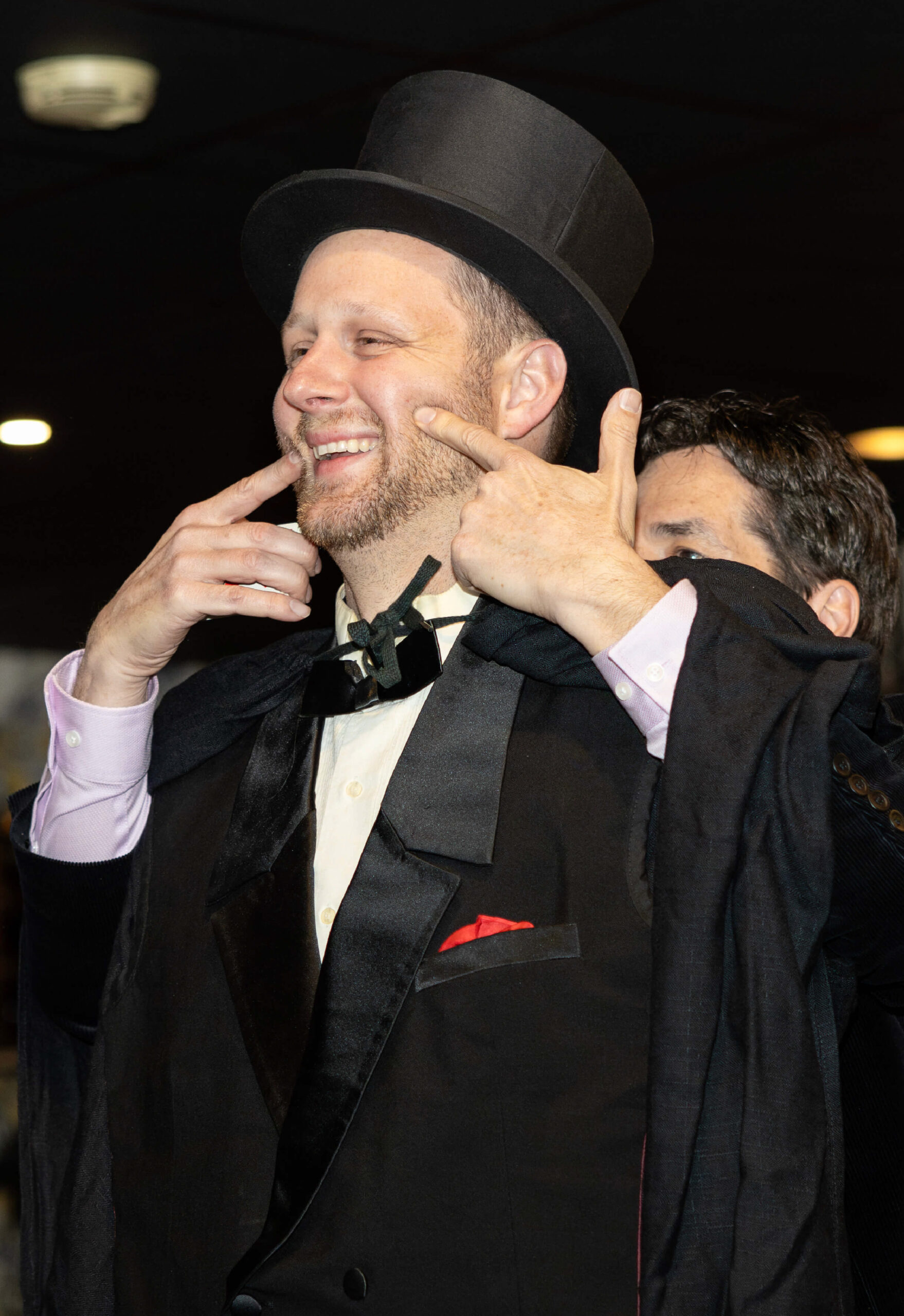 A smiling white man in a black top hat and tuxedo jacket. There is a partially obstructed second man standing behind him, holding his hands up to the first man's face.