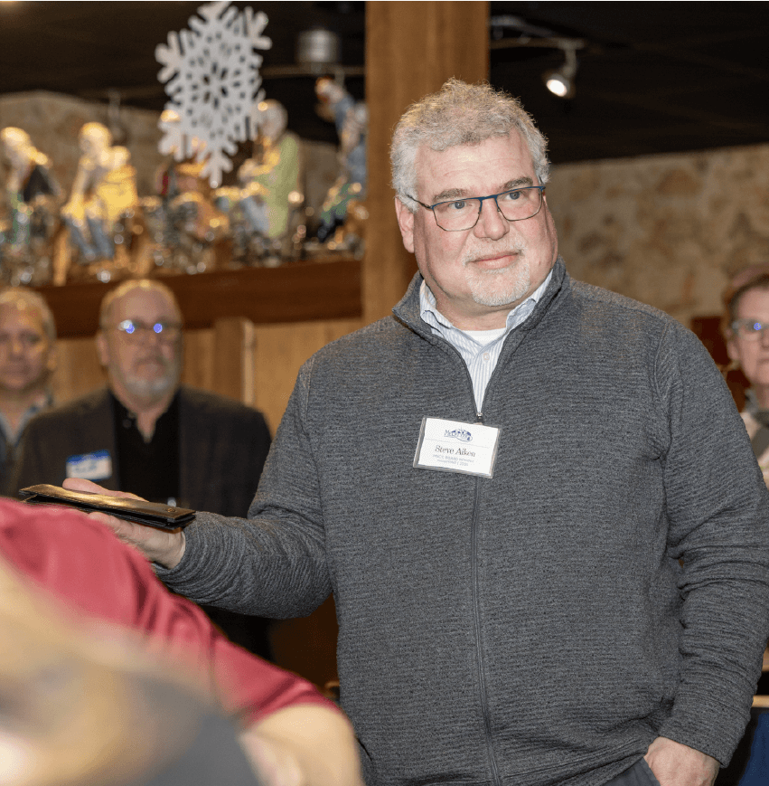 A man in a gray sweater is standing and holding a slim black leather folder in one outstretched palm.