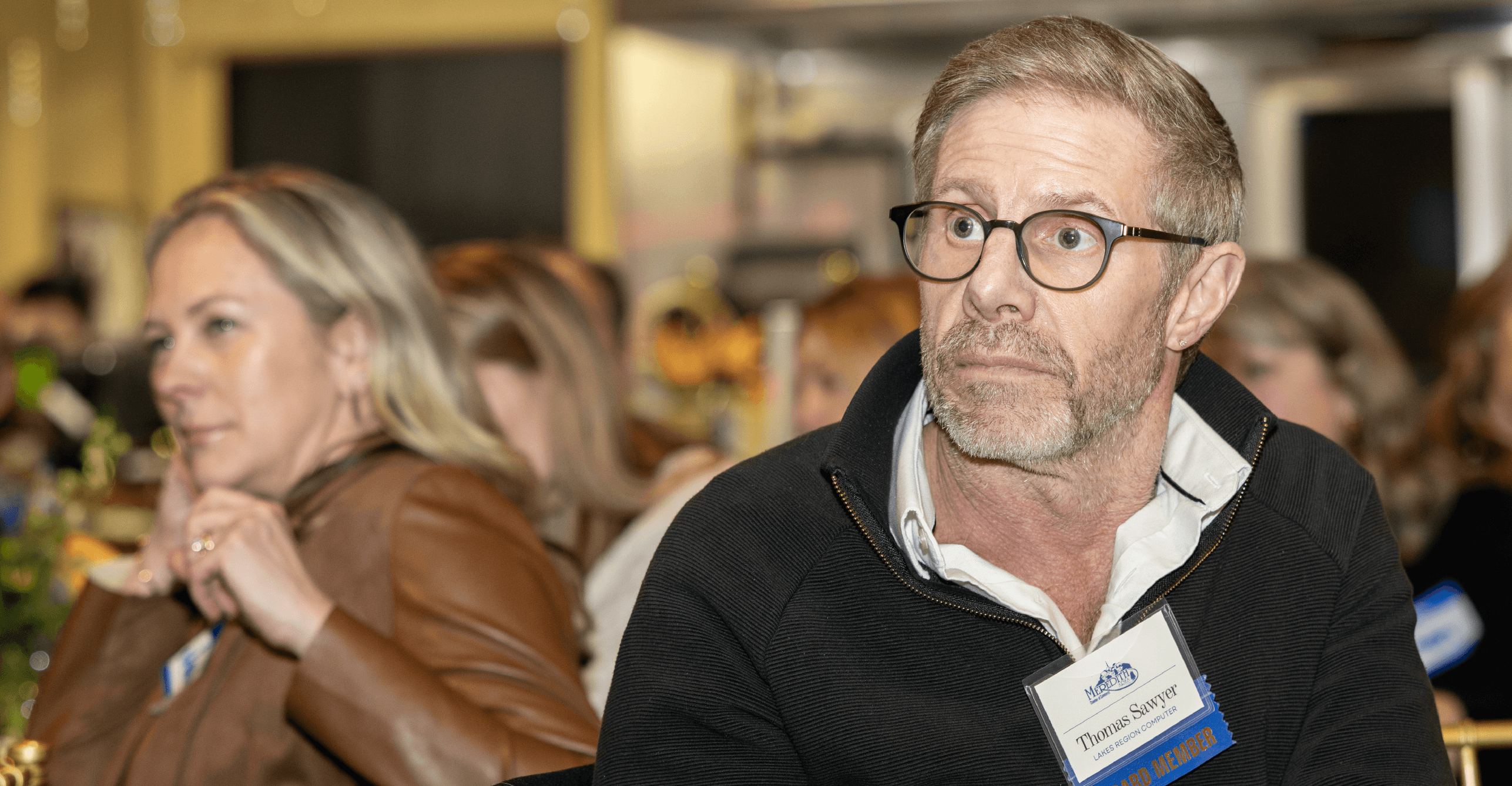 A white man in glasses and a name tag is in focus and looking to the left, while a room full of seated people are behind him out of focus.