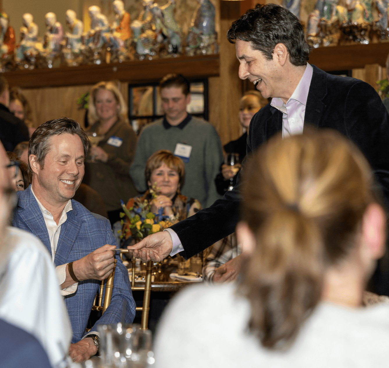 Two men smiling and facing each other surrounded by onlookers. One of the men is in the middle of performing a magic trick.